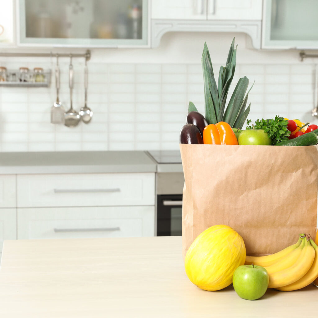 Paper shopping bag full of vegetables with fruits and juice on table in kitchen. Space for text